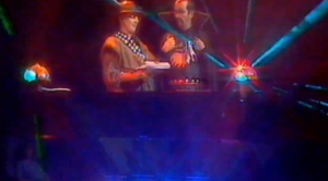 Man in cape and man in a hat talking in front of bright lights
