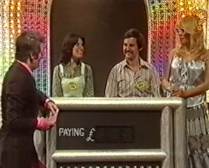 Host and contestants stood in front of a screen saying how much they are paying which is currently blank