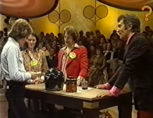 Three contestants stood around a table reading a card